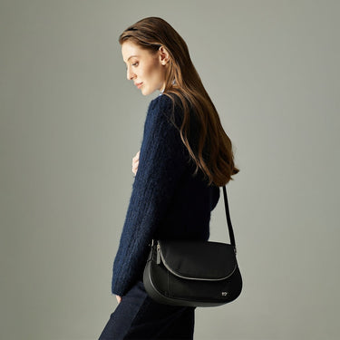 Model wearing a black leather shoulder bag with curved flap design, styled with a casual outfit.