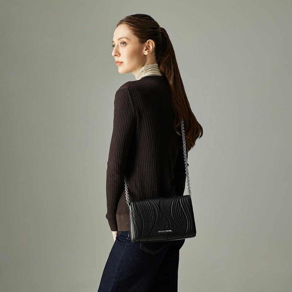 Model wearing a dark brown sweater and jeans, carrying a black striped crossbody bag with a silver chain